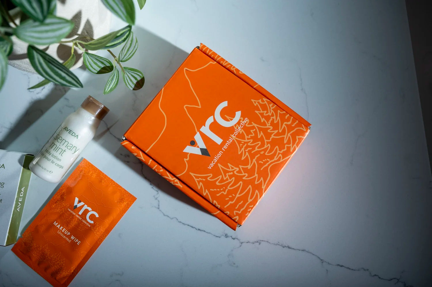 Orange VRC vacation rental box, makeup wipe, and Aveda lotion on a marble bathroom counter.