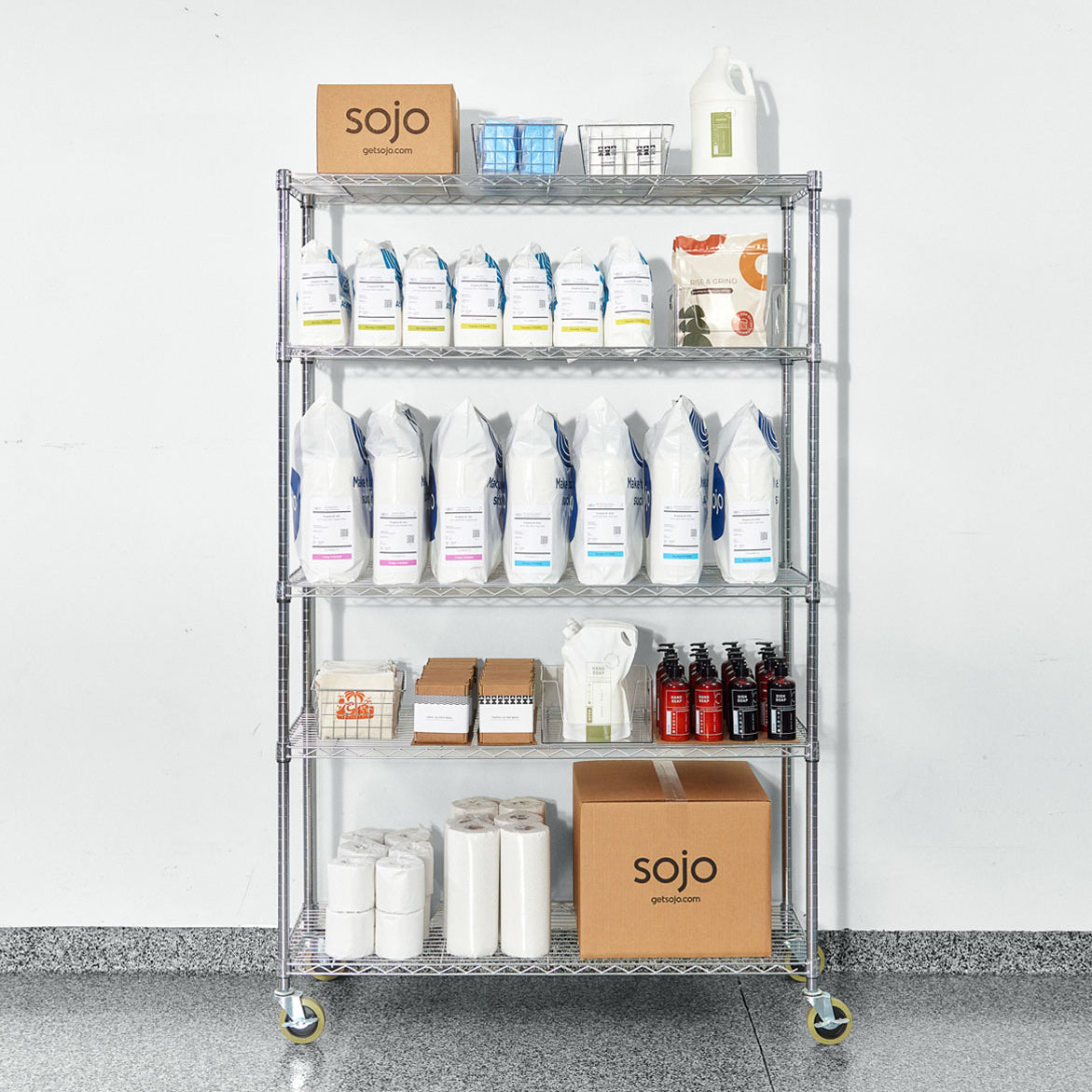 Metal shelving unit with Sojo cleaning products, boxes, bottles, and paper rolls against white wall.