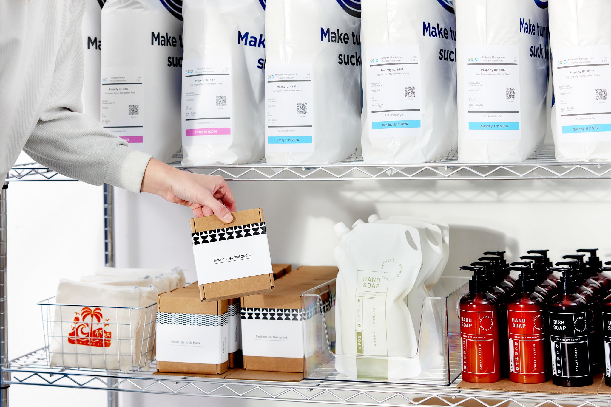 Hand reaching for boxed amenity kit on metal shelf with soaps and supply bags.