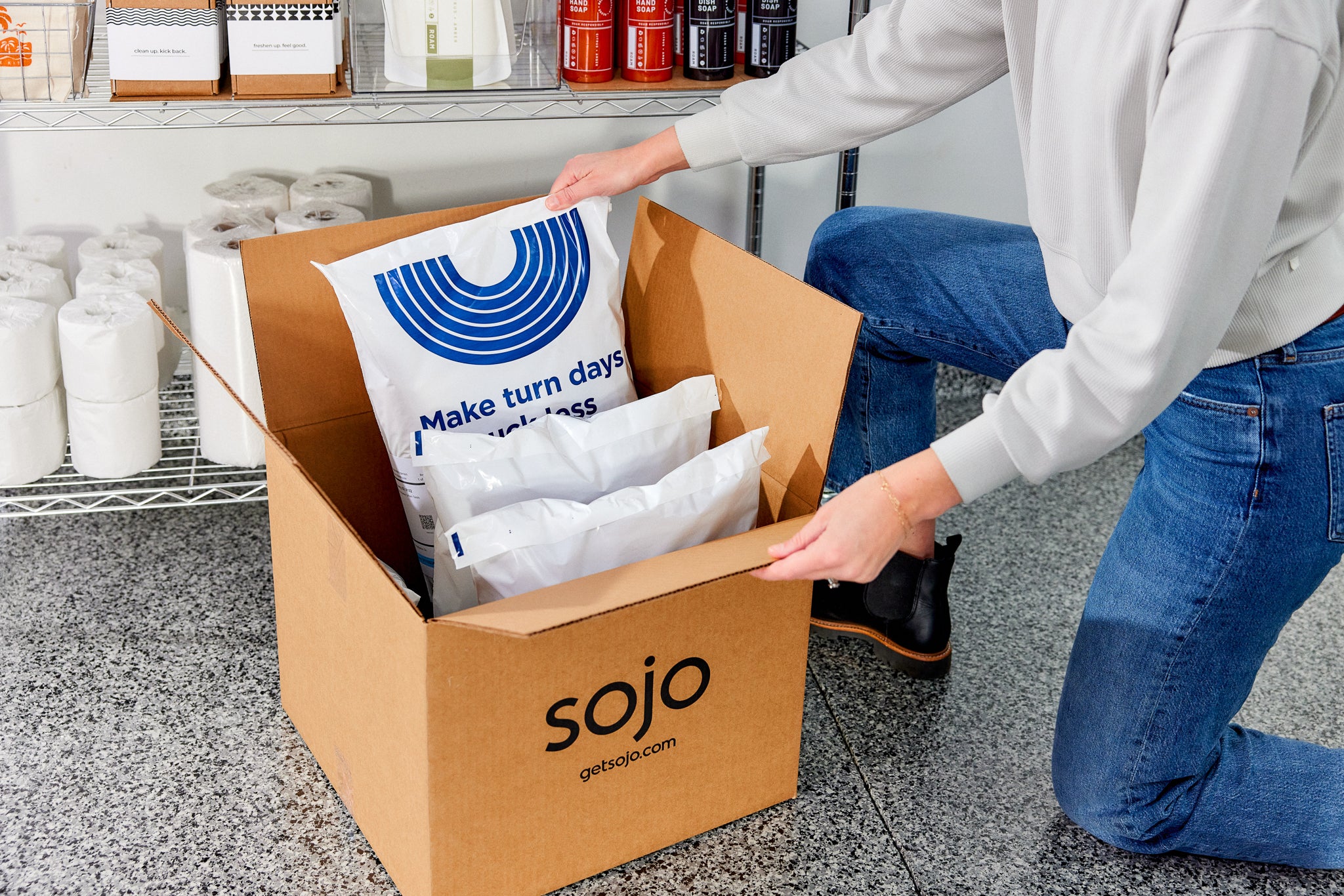 Person unpacking a Sojo box with branded packaging and toilet paper rolls on a shelf.
