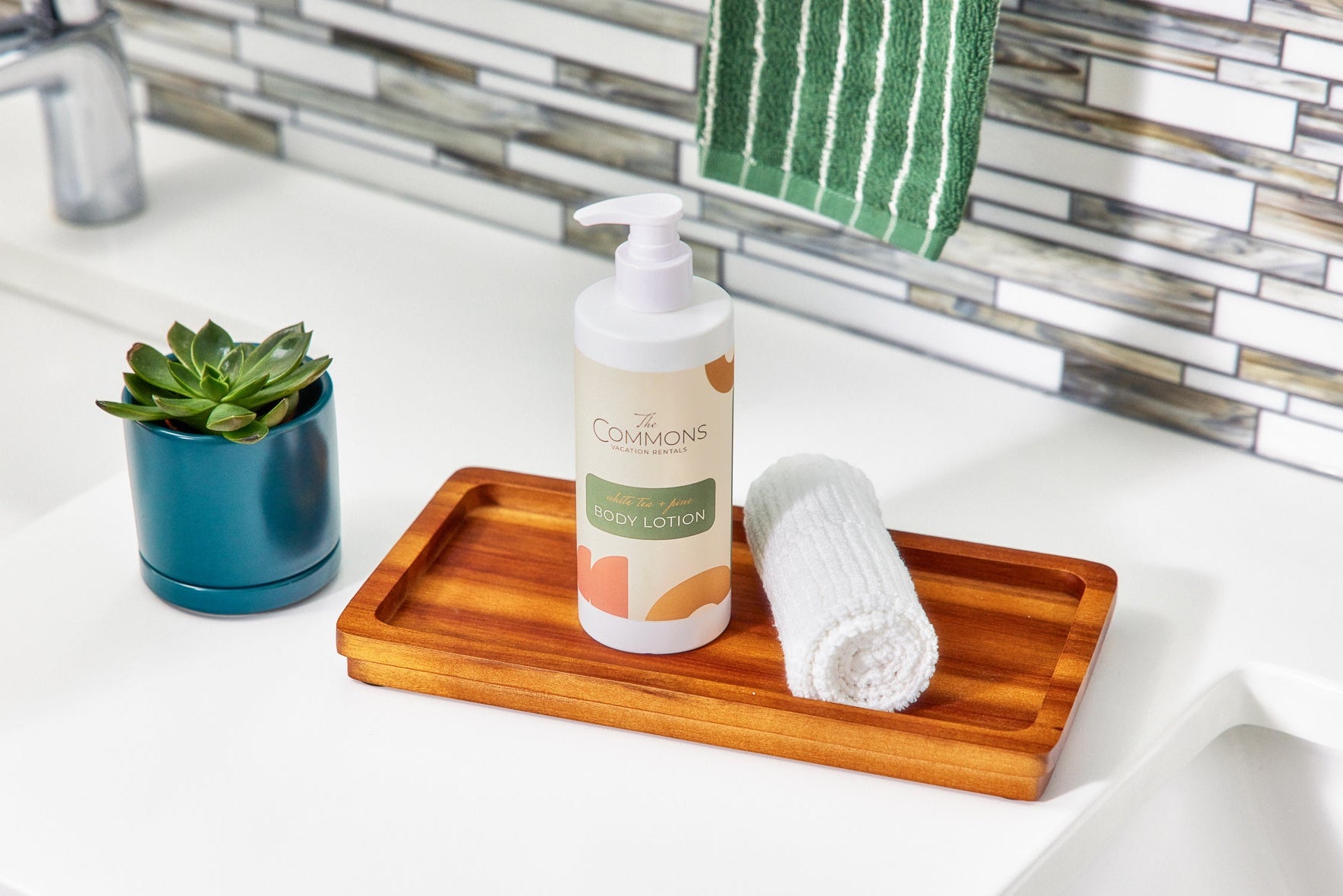 Bathroom counter with The Commons body lotion, rolled white towel on wood tray, and succulent plant.