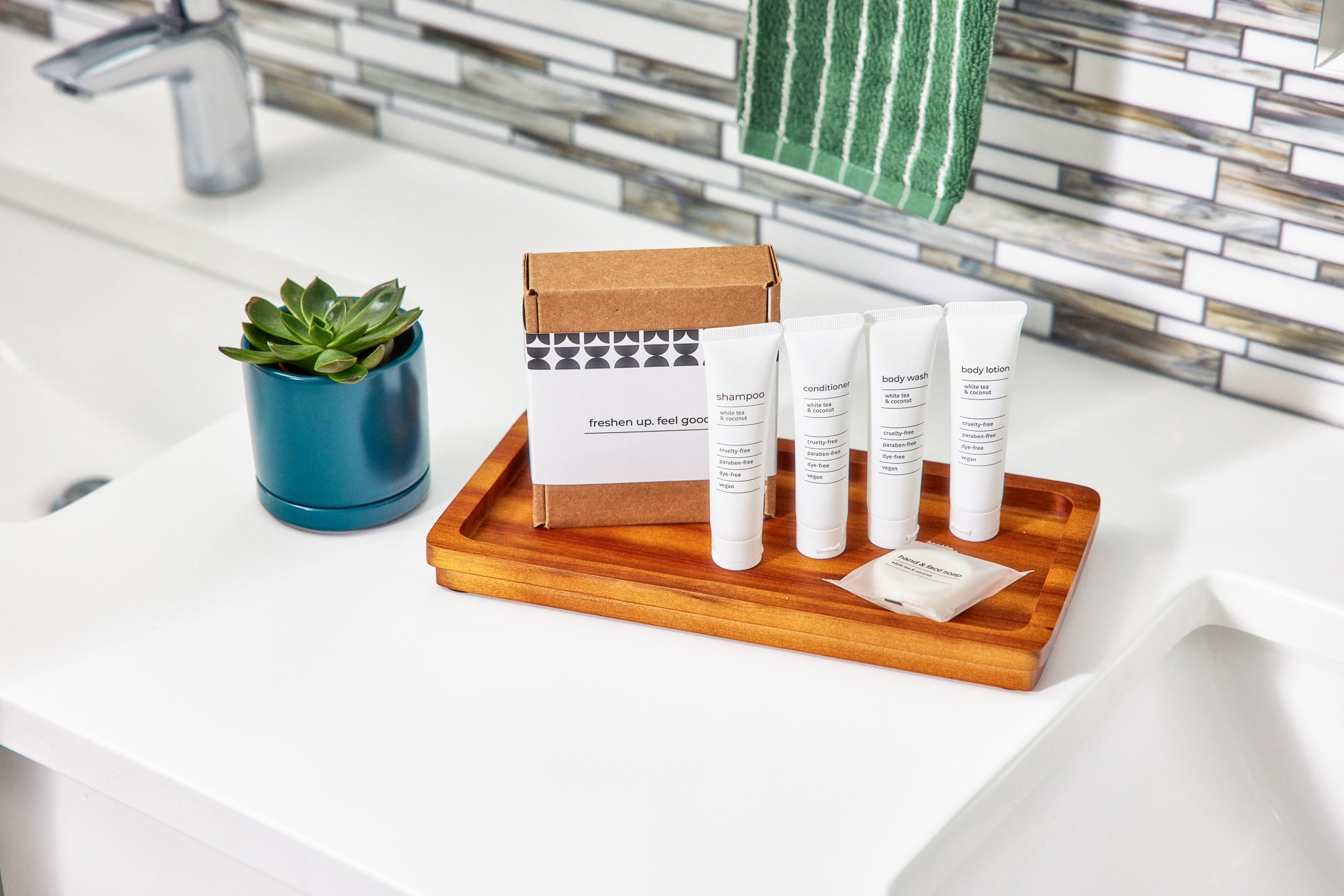 Travel toiletries kit with shampoo, conditioner, body wash, lotion on wood tray by bathroom sink.
