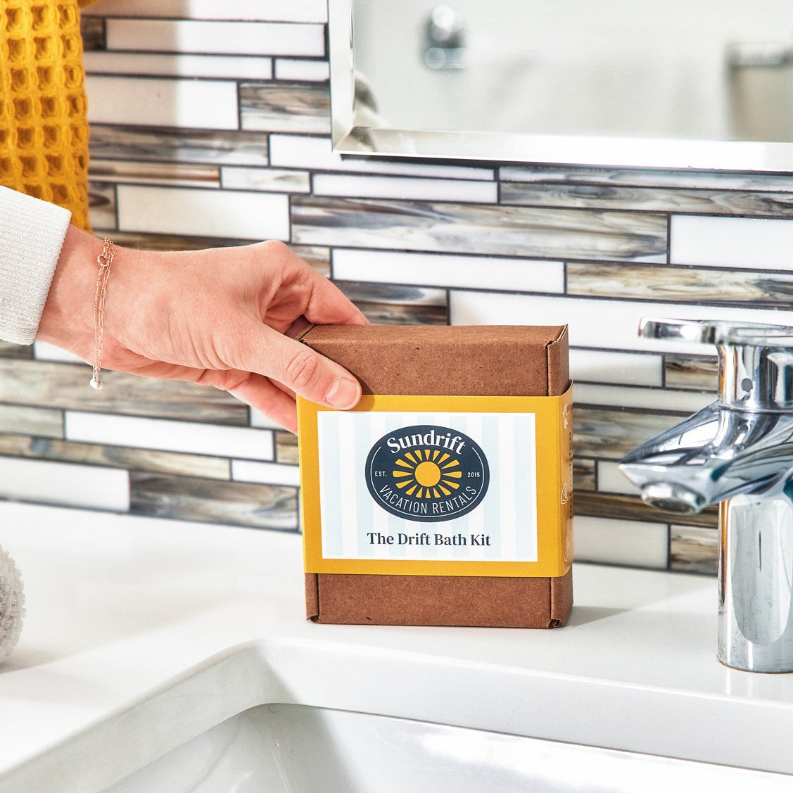 Hand placing Sundrift Vacation Rentals The Drift Bath Kit box on bathroom sink by faucet.