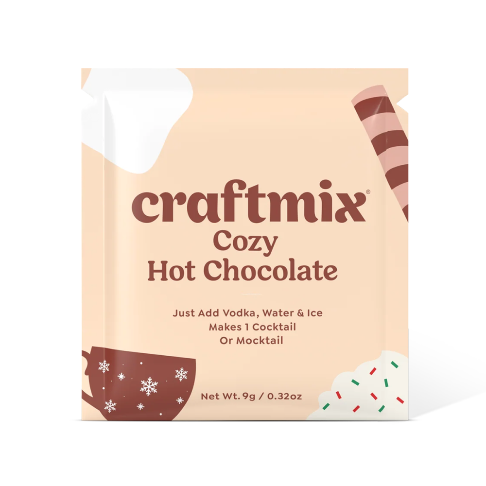 A single packet of craftmix Cozy Hot Chocolate cocktail mix on a clean white background. The packaging is a warm peach-beige color with dark brown text and holiday-themed illustrations, including a coffee cup with snowflakes, a striped drink straw, and white whipped cream with red and green sprinkles.
